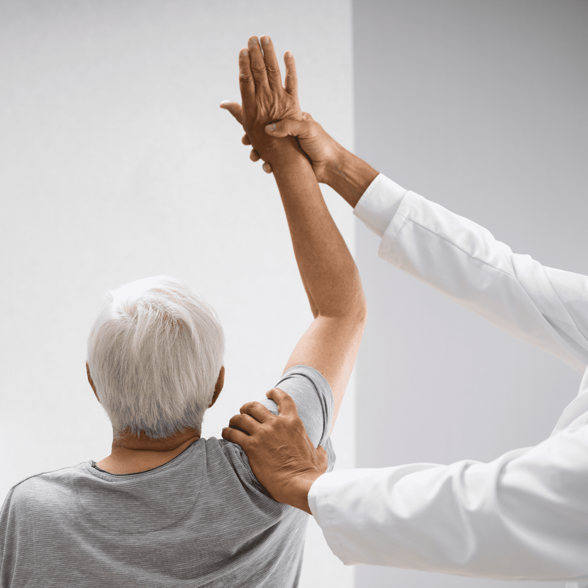 Clinician guides an older patient through a shoulder mobility exercise with arms raised during a physical therapy session.