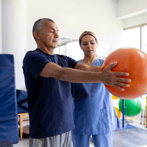 Older patient holds a large therapy ball during a rehab exercise as a clinician supervises and supports proper form. 