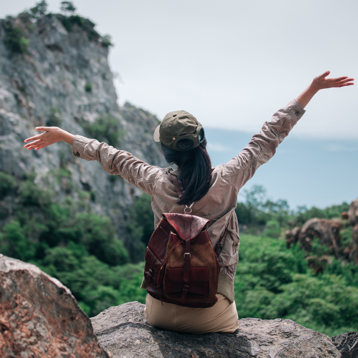 A woman sitting on a rock with arms outstretched overlooking a mountain valley, practicing travel therapy.