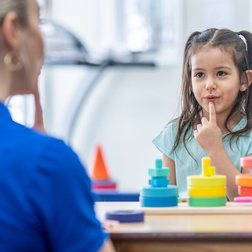 Child sits across from a clinician during a speech therapy session with colorful stacking toys on the table. 