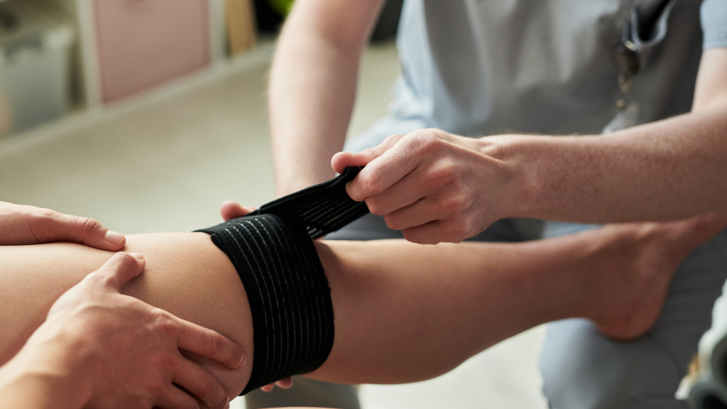 Sports Medicine Salary Trends and Specialized Care A sports medicine professional wrapping a patient's knee with a black compression bandage to treat an athletic injury.