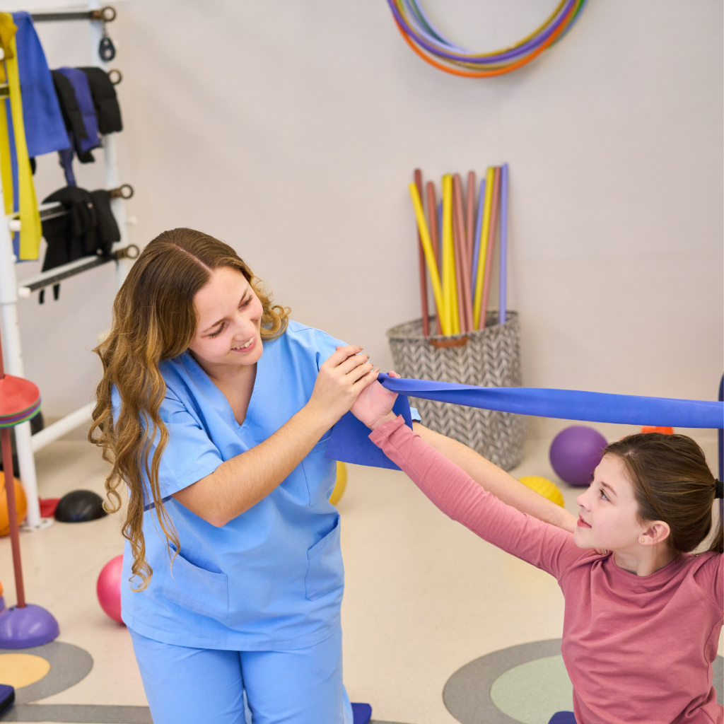 Physical therapist in blue scrubs guiding a child through resistance band stretching exercise in therapy clinic