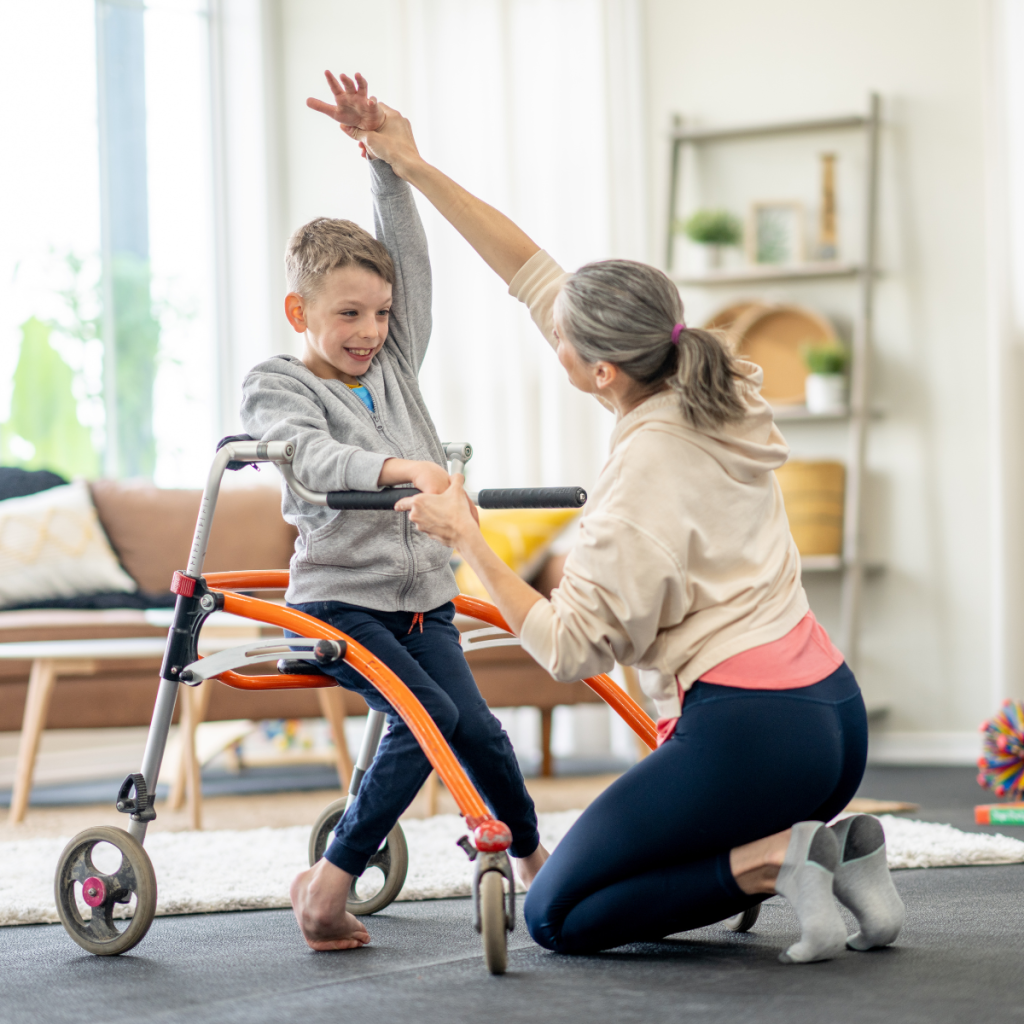 Physiotherapist kneeling beside young child learning to walk with a pediatric walker during rehabilitation session.