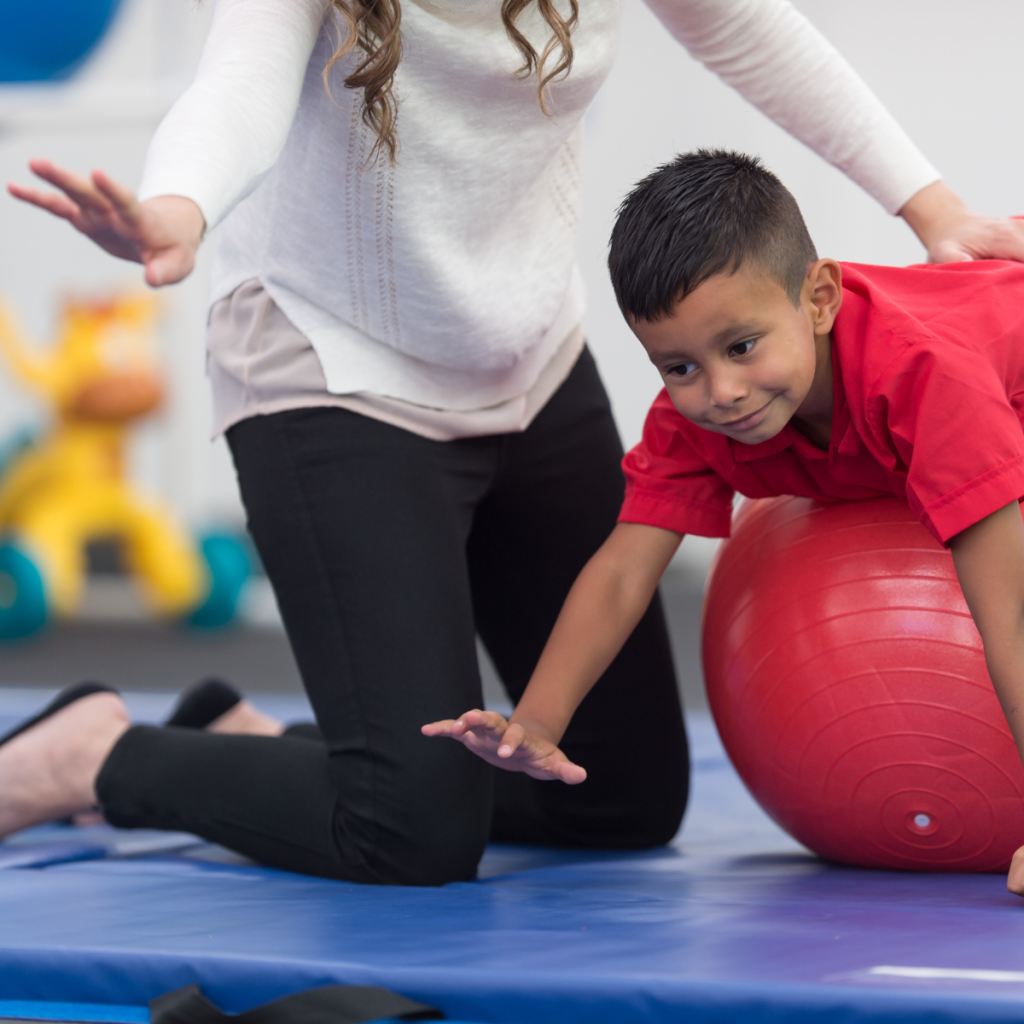 Pediatric Physical Therapist Working With Child on Exercise Ball in Clinic Pediatric physical therapist sitting on floor mat with smiling young boy balanced on red exercise ball during therapy session.