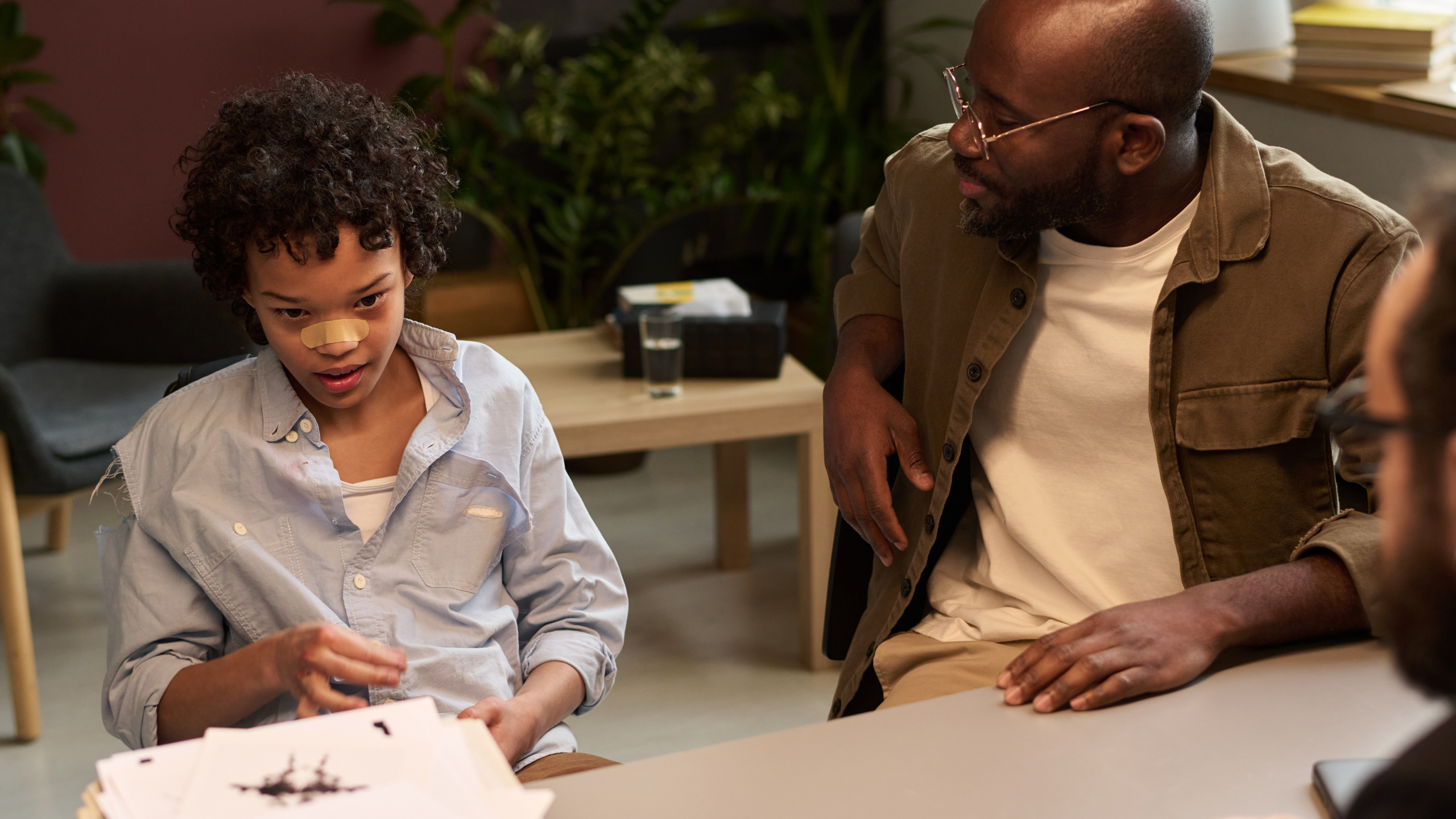 Young man looking at his naughty son in torn shirt answering questions of school psychologist during session in office.
