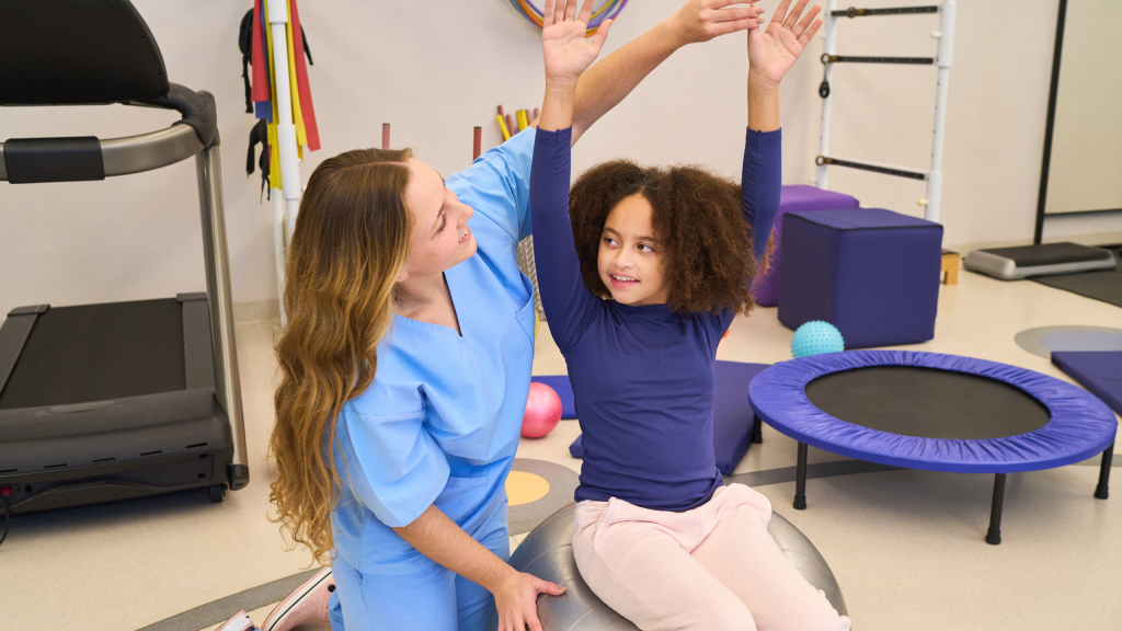 A pediatric rehabilitation session in a clinical gym where a clinician assists a young girl raised arms on a balance ball, promoting motor skills, coordination, and confidence in a supportive environment. 