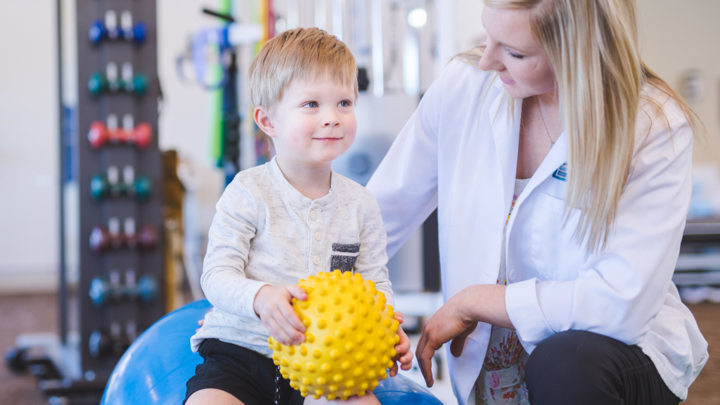 A blonde physical therapist works with a young boy in an open floor clinic. She is helping him do a fun exercise on a balance ball. He is looking at the camera and smiling while he holds a yellow exercise ball.  