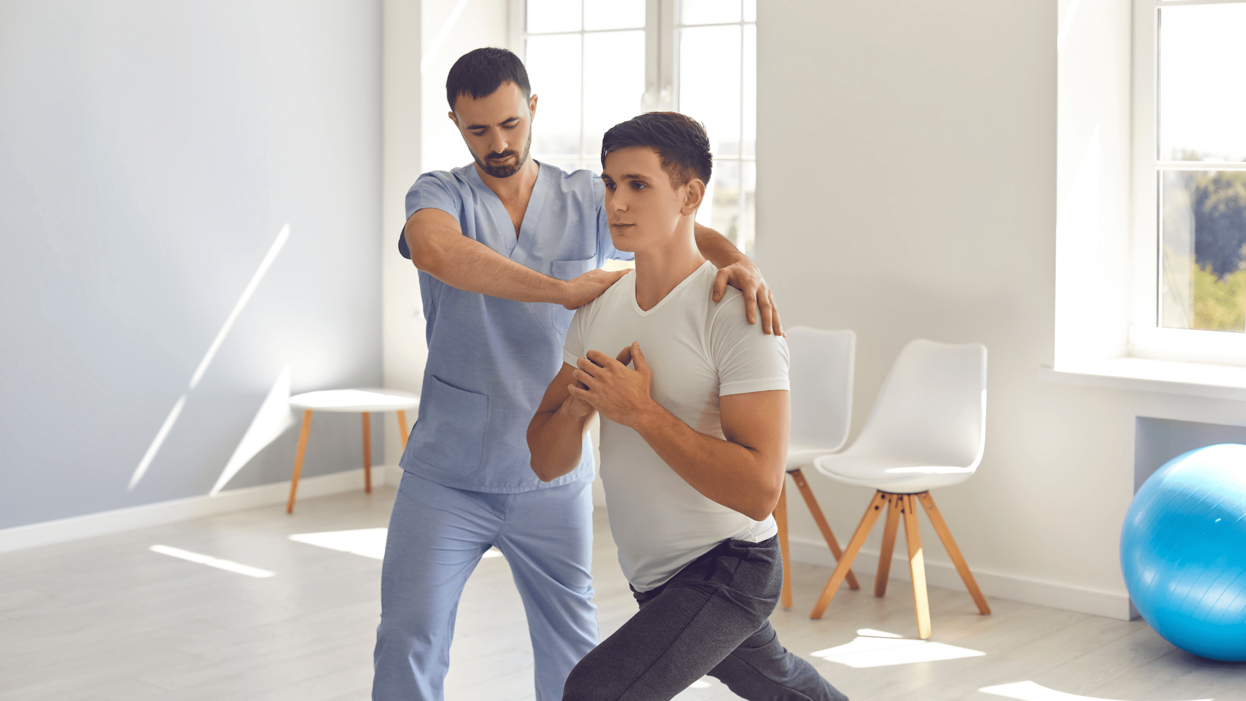 A physical therapist in blue scrubs guiding a male patient through a lunge exercise in a bright healthcare clinic.