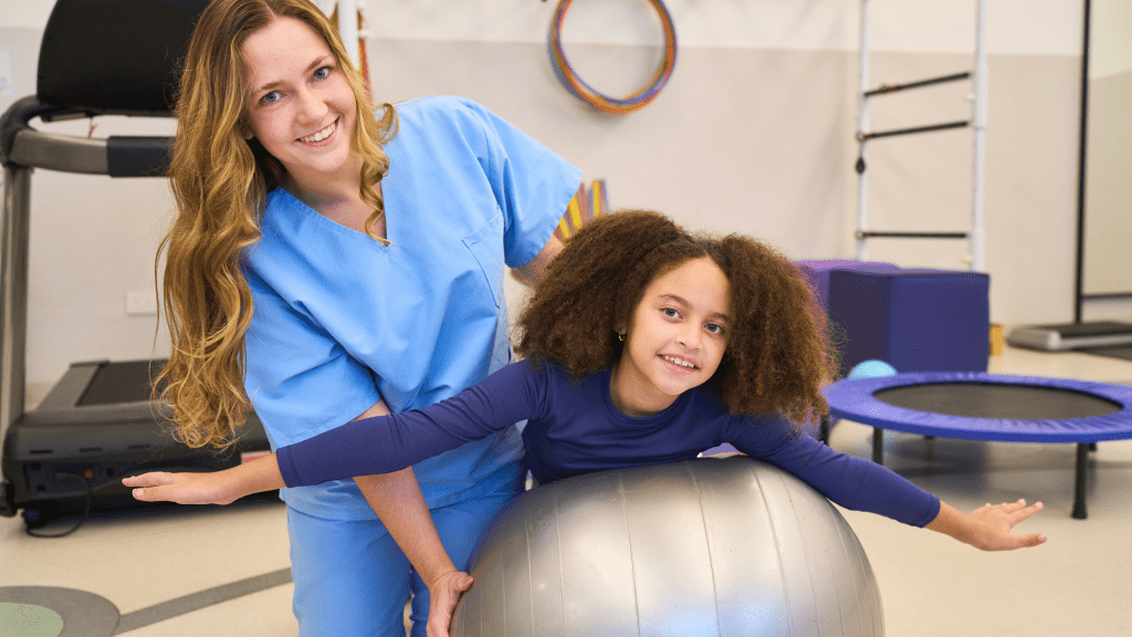 A smiling female physical therapist in blue scrubs assisting a young girl balancing on a large silver exercise ball in a colorful pediatric clinic. 