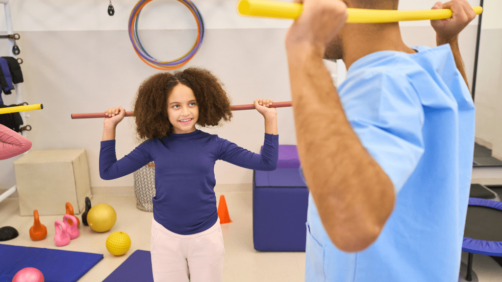 A pediatric physical therapist demonstrating a posture exercise with a yellow bar to a young girl holding a training wand in a rehab gym. 