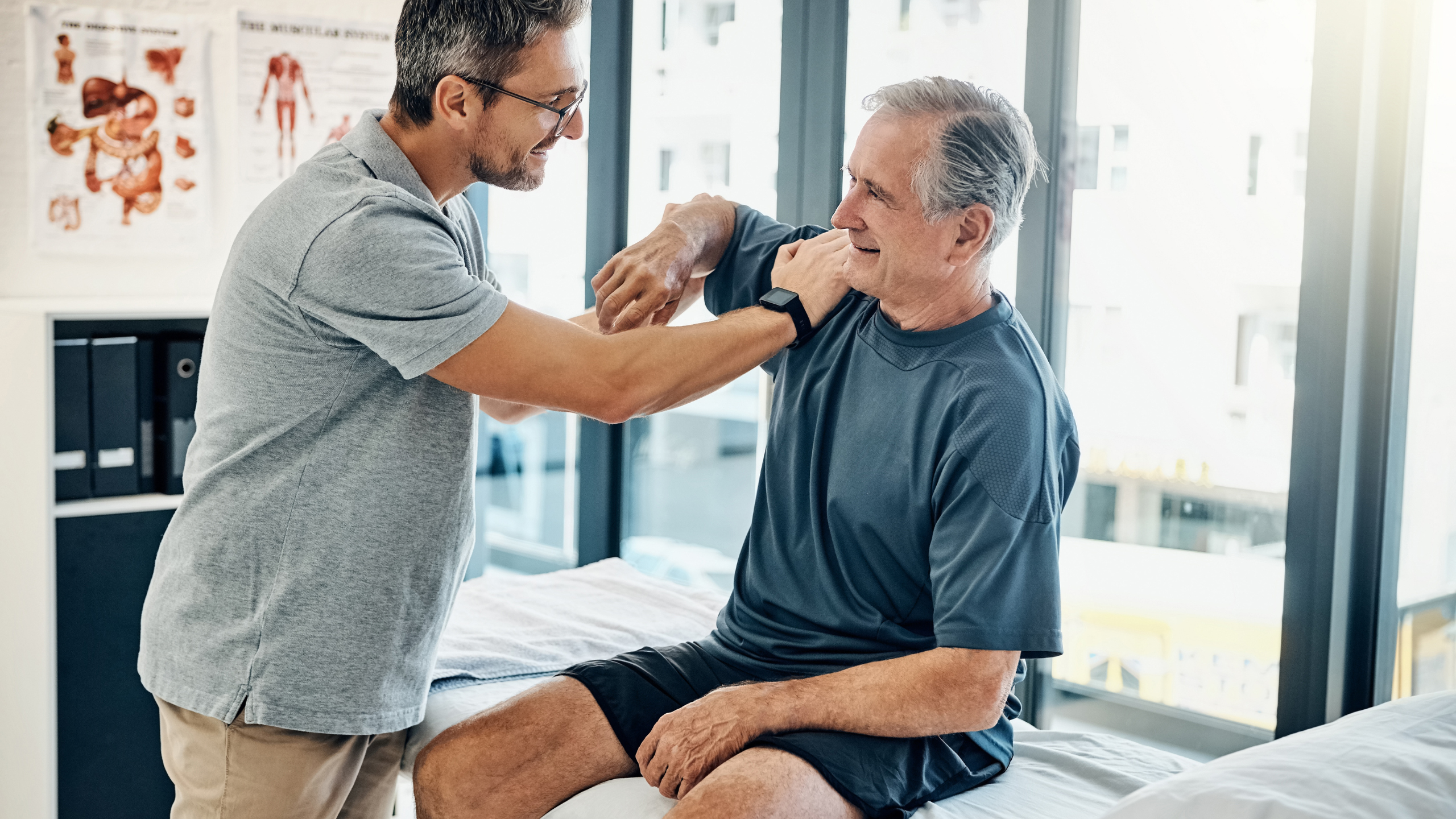 A male physiotherapist assessing the arm and elbow mobility of an elderly male patient in a bright rehab clinic.