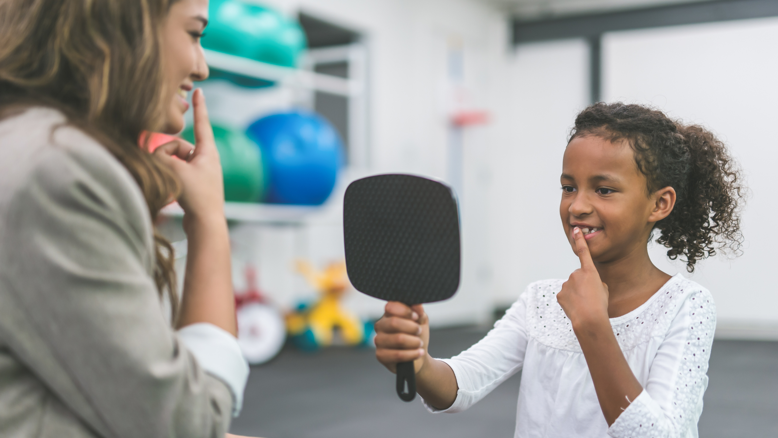 A female speech pathologist is teaching a child patient how to speak by having her touch her mouth and look into a mirror.
