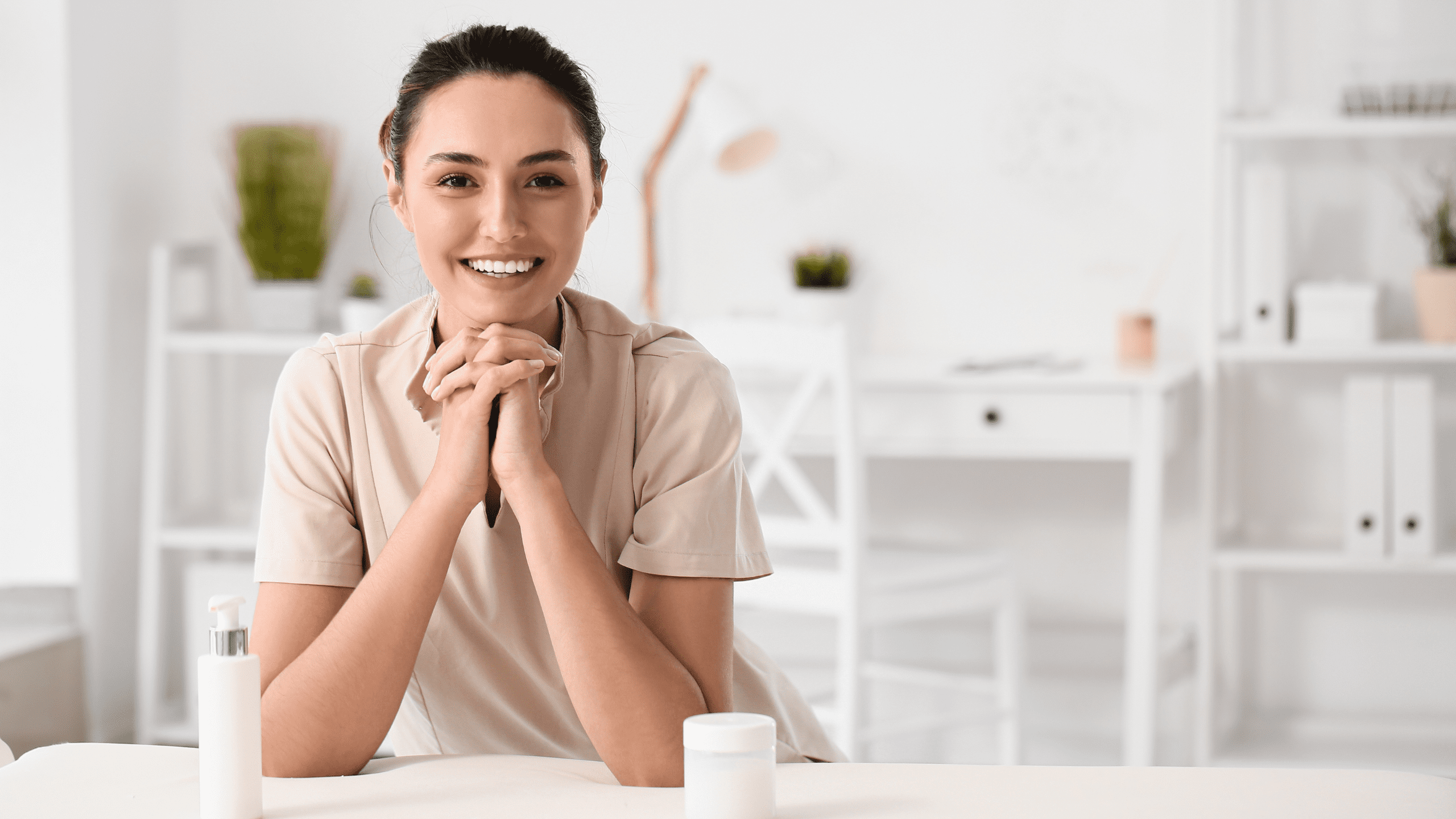 A smiling female therapist in a beige uniform leaning on a treatment table with lotion bottles in a bright, modern private practice office.