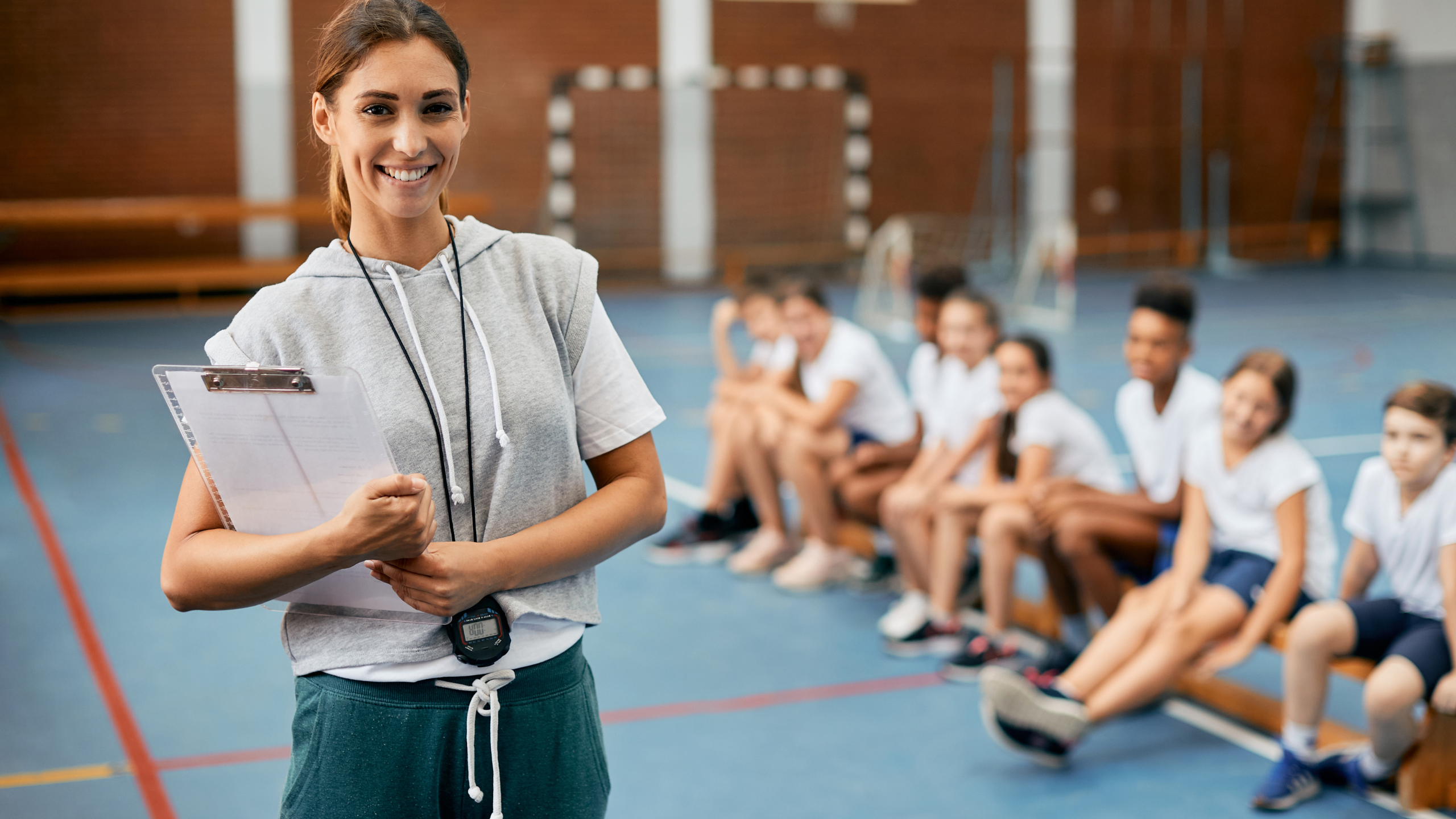 Young happy female coach looking at camera while having PE class with elementary students at school gym.