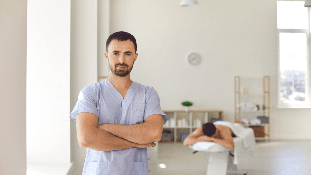 A confident male therapist in blue scrubs standing with arms crossed in a spacious healthcare clinic with a patient on a treatment table. 