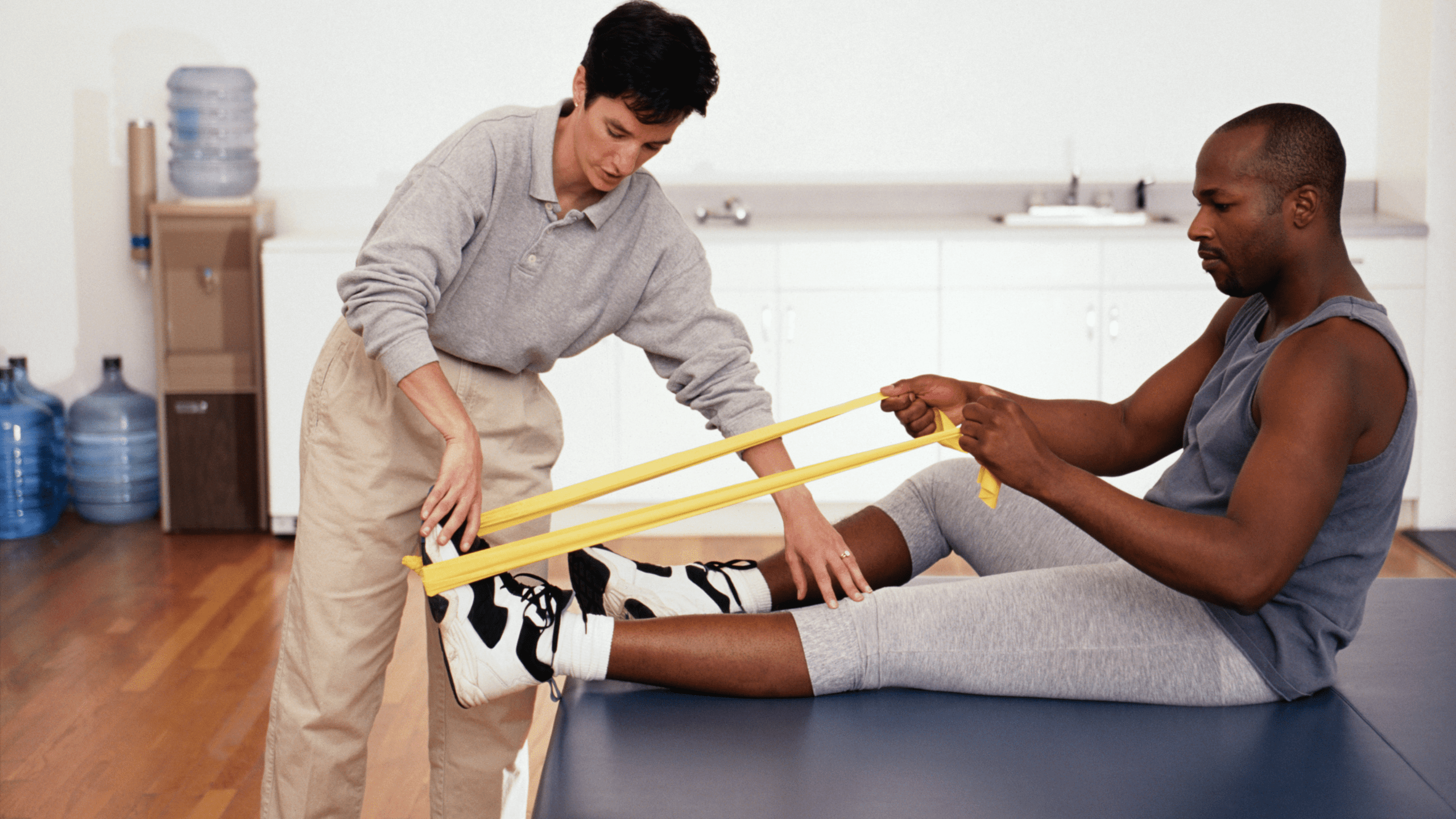 A female physical therapist assisting a male patient with a yellow resistance band leg exercise in a clinic setting.